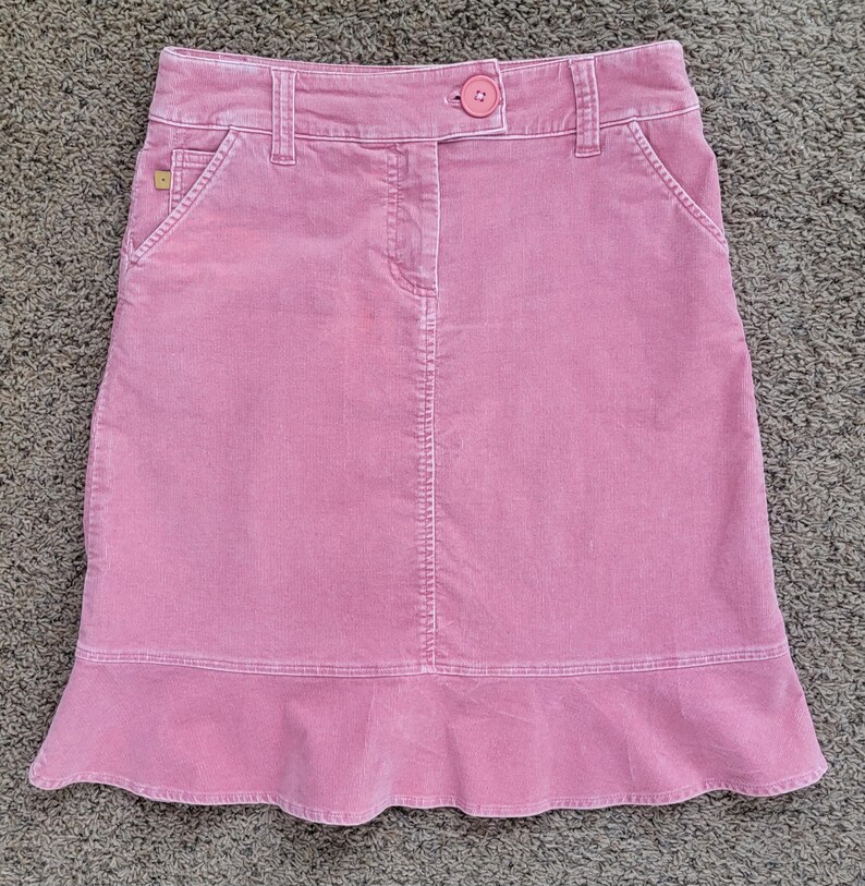 Op de afbeelding: Een roze corduroy rok met een knoopsluiting en zakken. De rok heeft een lichte A-lijn vorm en een gefranjerde zoom. De rok is gemaakt van een zachte, getextureerde stof.