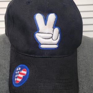 May include: Black baseball cap featuring a white peace sign with a blue outline on the front. The cap has a curved brim and a small patch with an American flag design on the side. The brim has red, white, and blue stripes.