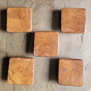 May include: Five square coasters with rounded corners, featuring a warm tan and brown mottled finish with dark speckles throughout. Each coaster has a smooth, flat surface and a uniform appearance, arranged in a pentagonal formation.