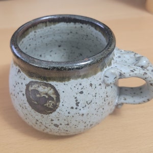 May include: A speckled ceramic mug with a rounded body and a unique handle design. The mug features a dark brown rim and a circular brown design on the side. The handle has a decorative, textured design.