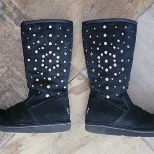 May include: Pair of black suede boots with a decorative pattern of white circular cutouts. The boots have a tall shaft, a flat sole, and the brand logo on the heel.