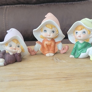 May include: Three ceramic elf figurines with blonde hair, blue eyes, and rosy cheeks. Each elf wears a different colored outfit: purple, orange, and green. They are sitting on a wooden surface, each with a whimsical hat.