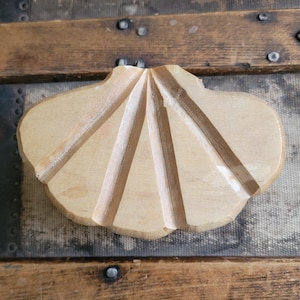 May include: A light brown, wooden, seashell-shaped soap dish. The dish has carved lines radiating from the center, creating a textured surface. The dish is set on a vintage wooden trunk.