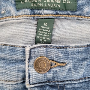 May include: Close-up of a pair of light blue denim jeans. The jeans feature a button closure with the words "LAUREN JEANS CO." and a green tag with the text "LRL LAUREN JEANS CO. RALPH LAUREN". The tag also includes the size 10 and fabric information.