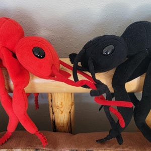 May include: Two plush toy ants, one red and one black, are positioned on a wooden bench. The red ant is on the left, and the black ant is on the right. Both have multiple legs, antennae, and large, black eyes.