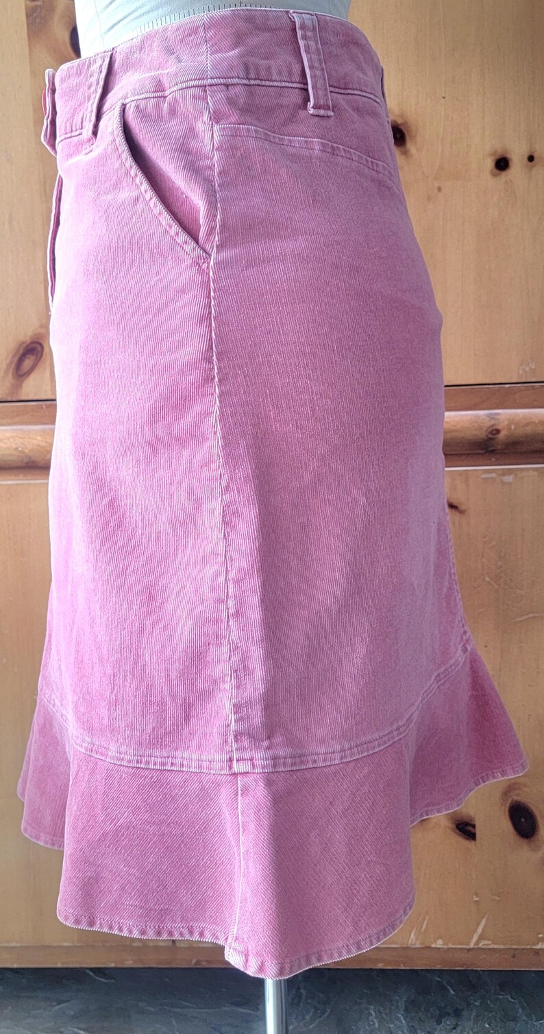 Op de afbeelding: Een roze corduroy rok met een uitlopende zoom. De rok heeft een klassiek ontwerp met vijf zakken, waaronder een voorzak. De stof heeft een verticale ribtextuur. De rok is effen roze van kleur.