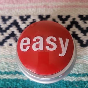 May include: A red button with the word "easy" in white, set on a silver base. The button is round and has a glossy finish. The background is a colorful patterned textile.