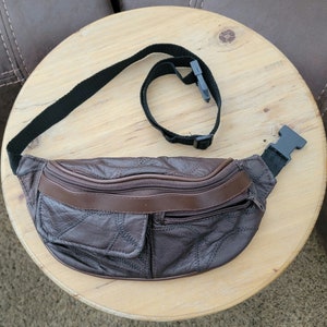May include: A brown leather fanny pack with a black adjustable strap and buckle. The fanny pack has a quilted design, a front pocket, and two zippered compartments. The fanny pack is sitting on a wooden table.