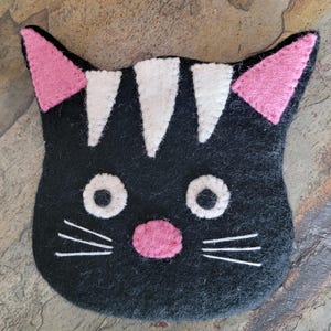 May include: Black felt cat-shaped coaster with pink inner ears, a pink nose, white whiskers, and white stripes on the forehead. The coaster is designed to resemble a cat's face and is a decorative item.