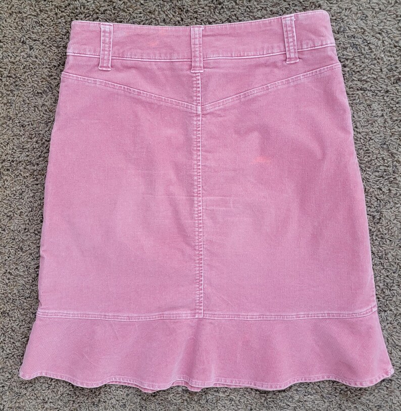Op de afbeelding: Een stoffen roze corduroy rok met een uitlopende zoom. De rok heeft een klassiek ontwerp met vijf zakken, een tailleband en riemlussen. De achterkant heeft een rugstuk en een verticale naad in het midden.