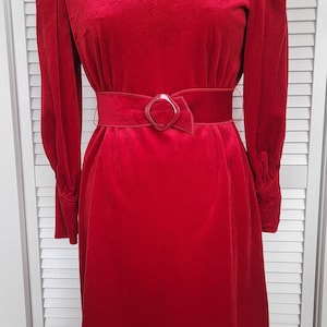May include: A vibrant red velvet dress with long sleeves and a matching belt. The dress features a boat neckline and a decorative buckle on the belt. A silver necklace adds a touch of elegance to the ensemble. The dress is displayed on a mannequin.