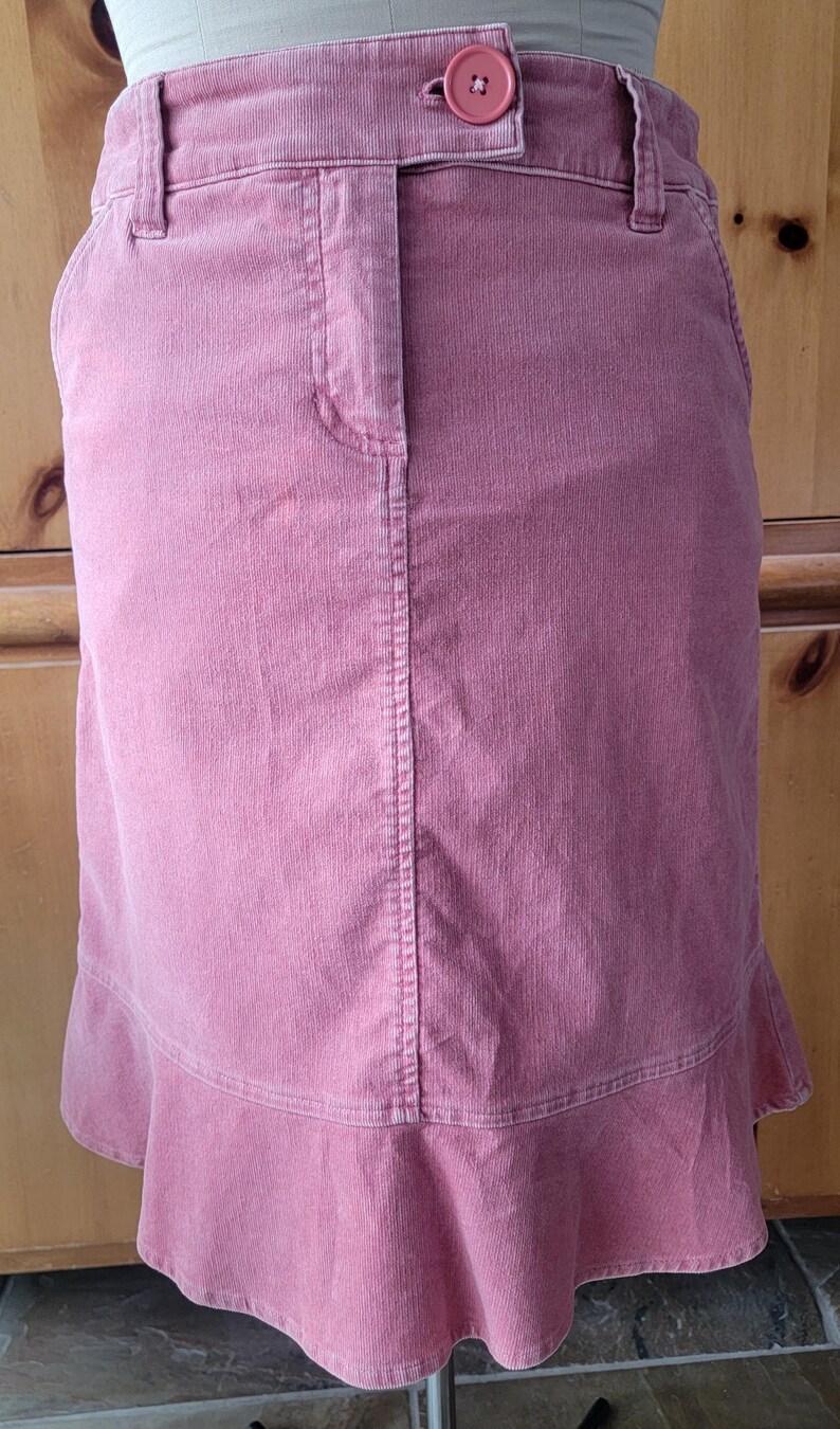 Op de afbeelding: Een stoffen rok in stoffig roze met een knoopsluiting en een gefranjerde zoom. De rok heeft een klassiek ontwerp met vijf zakken en een getailleerd silhouet. De rok is gemaakt van een zachte, getextureerde corduroy stof.