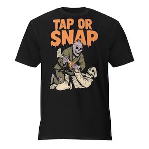 May include: Black t-shirt featuring a graphic of two skeleton figures in a martial arts pose. The text "TAP OR SNAP" is in orange above the figures. The skeletons are in green and beige attire.