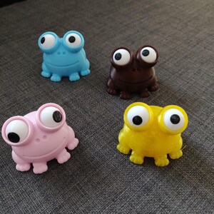 Puede incluir: Cuatro pequeñas figuras de rana en azul, marrón, rosa y amarillo. Cada rana presenta grandes ojos blancos saltones con pupilas negras, un cuerpo redondeado y liso, y cuatro pies pequeños. Las ranas están dispuestas en un grupo.