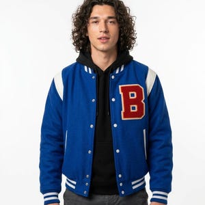 May include: A blue varsity jacket with white sleeves and striped ribbed cuffs, collar, and hem. The jacket features a large red letter B patch with a white border on the left chest, silver snap buttons, and is worn over a black hooded sweatshirt.