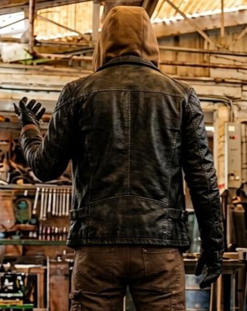 May include: A dark charcoal gray jacket with a quilted shoulder texture and a back seam, worn over a tan hooded sweatshirt. The jacket features long sleeves, a straight hemline, and is paired with brown pants and black gloves, viewed from the back.