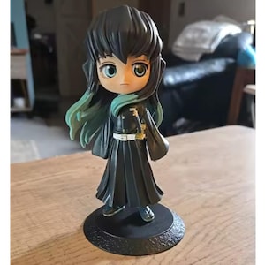 May include: A small figurine of a character with black hair and teal highlights, wearing a black outfit with a white belt and a small sword. The figure has large, expressive eyes and stands on a black base.