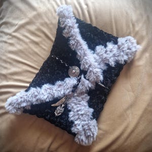 May include: Black decorative pillow with gray faux fur trim. The pillow features a silver-colored metal clasp with a decorative pendant and chain. The pillow is on a tan surface.