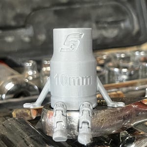 以下が含まれることがあります： レンチの上に座っている、腕と脚が付いた灰色の10mmソケット。ソケットにはSnap-onのロゴと「10mm」の数字が印刷されています。このソケットはノベルティグッズです。