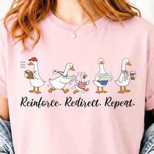 May include: A pale pink t-shirt with a cartoon of five geese in different poses. The geese are wearing a red cap, carrying a clipboard, running with a data transfer, wearing a striped shirt, and holding a coffee cup. The text "Reinforce. Redirect. Repeat." is below the geese.