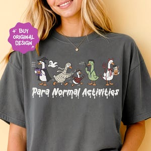 Peut inclure: T-shirt gris foncé avec un motif sur le thème d'Halloween. Le dessin présente des canards de dessins animés déguisés, avec l'inscription "Para Normal Activities". Le t-shirt a une coupe ample.