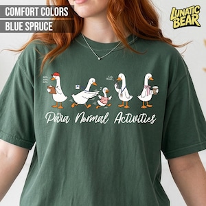 May include: Blue spruce-colored t-shirt featuring a whimsical design of cartoon geese engaged in various activities. The phrase "Para Normal Activities" is written in a script font below the geese. The shirt also has the text "Comfort Colors" and "Blue Spruce".