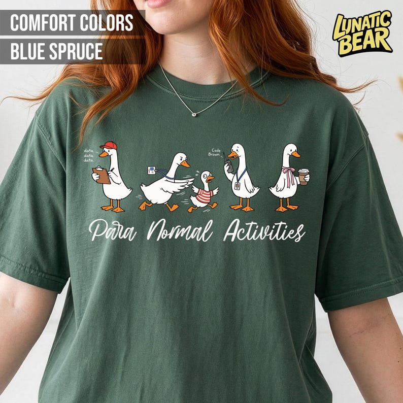 May include: Blue spruce-colored t-shirt featuring a whimsical design of cartoon geese engaged in various activities. The phrase "Para Normal Activities" is written in a script font below the geese. The shirt also has the text "Comfort Colors" and "Blue Spruce".