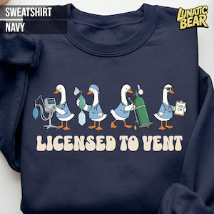 Könnte beinhalten: Marineblaues Sweatshirt mit einer Grafik von Cartoon-Gänsen in medizinischer Kleidung. Die Gänse halten medizinische Geräte, darunter steht der Text "LICENSED TO VENT". Das Sweatshirt hat auch den Text "SWEATSHIRT NAVY" in der oberen linken Ecke.