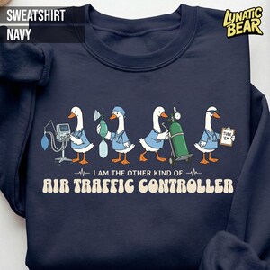 May include: Navy blue sweatshirt with a whimsical design featuring cartoon geese dressed as medical professionals. The geese are holding medical equipment and the text reads "I AM THE OTHER KIND OF AIR TRAFFIC CONTROLLER."