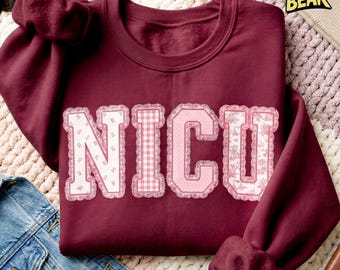 May include: A maroon sweatshirt featuring the word "NICU" in pink, with a decorative floral and gingham pattern. The garment is folded on a patterned surface, with a denim jacket and sunglasses visible.