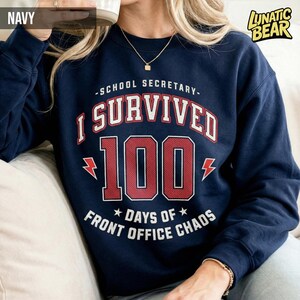 May include: Navy blue sweatshirt with the text "-SCHOOL SECRETARY- I SURVIVED 100 DAYS OF FRONT OFFICE CHAOS" in red and white lettering. The number 100 is in a bold, red font. The sweatshirt has a crew neck and long sleeves.