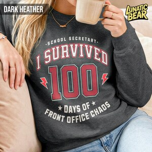 May include: Dark heather gray sweatshirt with the text "SCHOOL SECRETARY - I SURVIVED 100 DAYS OF FRONT OFFICE CHAOS" in red and white. The sweatshirt has a crew neck and long sleeves. The brand logo "LUNATIC BEAR" is in the upper right corner.