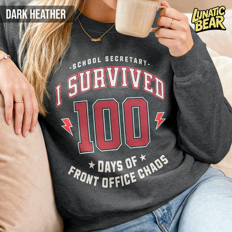 May include: Dark heather gray sweatshirt with the text "SCHOOL SECRETARY - I SURVIVED 100 DAYS OF FRONT OFFICE CHAOS" in red and white. The sweatshirt has a crew neck and long sleeves. The brand logo "LUNATIC BEAR" is in the upper right corner.