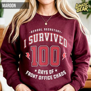 May include: Maroon sweatshirt with the text "SCHOOL SECRETARY - I SURVIVED 100 DAYS OF FRONT OFFICE CHAOS" in white and red. The number 100 is in a large, bold font with lightning bolt graphics on either side. The sweatshirt has a crew neck.
