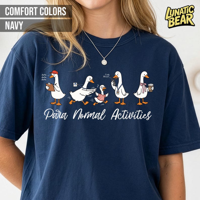 May include: Navy blue t-shirt with a graphic of cartoon geese engaged in various activities. The text "Para Normal Activities" is printed below the geese. The shirt also features the text "Comfort Colors Navy" and a "Lunatic Bear" logo.