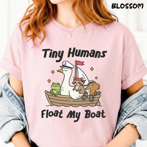 Peut inclure: T-shirt rose clair avec l'inscription "Tiny Humans Float My Boat". Le motif représente un bateau de dessin animé avec une oie portant un chapeau de capitaine, une grenouille, un raton laveur et un capybara. Le mot "Blossom" est imprimé en haut à droite.