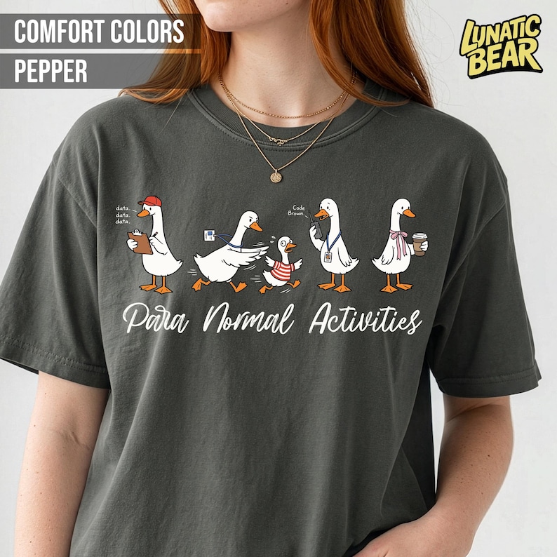 May include: Dark grey t-shirt with a whimsical illustration of cartoon geese engaged in various activities. The text "Para Normal Activities" is printed below the geese. The shirt also features the text "Comfort Colors Pepper" and the "Lunatic Bear" logo.