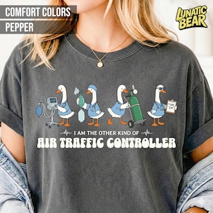 May include: Dark grey t-shirt with a graphic of four cartoon geese in medical attire. The geese are holding medical equipment. Text on the shirt reads "I AM THE OTHER KIND OF AIR TRAFFIC CONTROLLER."