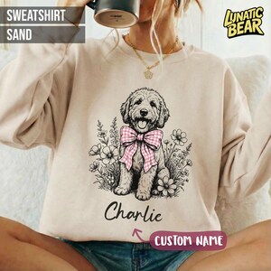 Op de afbeelding: Zandkleurige sweatshirt met een afbeelding van een hond met een roze en wit geblokte strik, omringd door bloemen. De naam "Charlie" staat onder de hond, met "CUSTOM NAME" eronder. De linkerbovenhoek leest "SWEATSHIRT SAND".