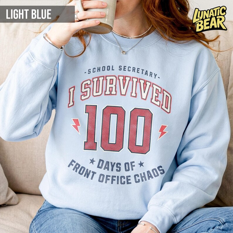 May include: Light blue sweatshirt with the text "-SCHOOL SECRETARY- I SURVIVED 100 DAYS OF FRONT OFFICE CHAOS" in red and white. The sweatshirt has long sleeves and a crew neck. The brand logo "LUNATIC BEAR" is in the upper right corner.