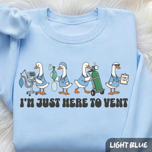 May include: A light blue sweatshirt featuring a graphic of four cartoon ducks dressed as medical professionals with equipment. The text below the ducks reads, "I'M JUST HERE TO VENT." The ducks are wearing blue scrubs and stethoscopes.