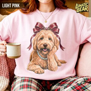 May include: Light pink sweatshirt featuring a detailed illustration of a golden doodle dog wearing a pearl necklace and a burgundy bow. The text "LIGHT PINK" is at the top left, and "LUNATIC BEAR" is at the top right.