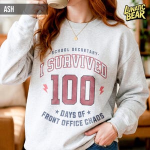 May include: Ash gray sweatshirt with the text "SCHOOL SECRETARY - I SURVIVED 100 DAYS OF FRONT OFFICE CHAOS" in red and white. The sweatshirt has a crew neck and long sleeves. The brand logo "LUNATIC BEAR" is in the upper right corner.