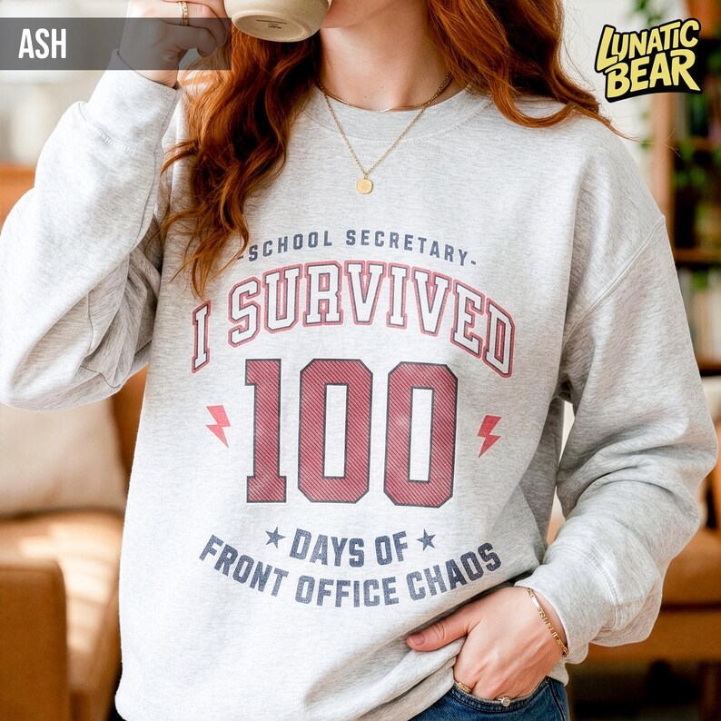 May include: Ash gray sweatshirt with the text "SCHOOL SECRETARY - I SURVIVED 100 DAYS OF FRONT OFFICE CHAOS" in red and white. The sweatshirt has a crew neck and long sleeves. The brand logo "LUNATIC BEAR" is in the upper right corner.