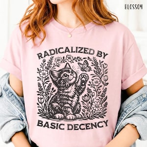 May include: A light pink t-shirt with the text "RADICALIZED BY BASIC DECENCY" in black. The graphic features a black and white illustration of a kitten reaching for a butterfly, surrounded by flowers and foliage. The word "BLOSSOM" is printed at the top.