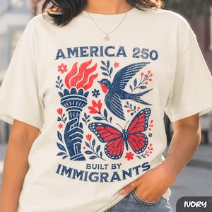 以下が含まれることがあります： アイボリー色のTシャツ。愛国的なデザインで、トーチ、鳥、蝶が赤と青で描かれ、「AMERICA 250 BUILT BY IMMIGRANTS」の文字が入っています。