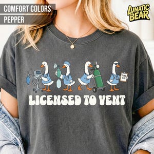 May include: Dark gray t-shirt with the text "LICENSED TO VENT" and a graphic of cartoon ducks dressed as medical professionals. The ducks are depicted with medical equipment, including a ventilator, oxygen tank, and nebulizer.
