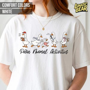 May include: White t-shirt with a graphic of cartoon geese engaged in various activities. The geese are depicted with accessories like a clipboard, a messenger bag, and a coffee cup. The text "Para Normal Activities" is printed below the geese.