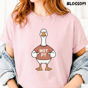 Peut inclure: T-shirt rose clair avec un dessin d'oie portant un t-shirt marron avec le texte "NOT PT". L'oie est blanche avec un bec et des pattes oranges. Le mot "BLOSSOM" est imprimé en haut à droite.
