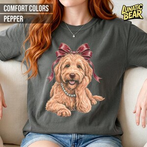 May include: Dark grey t-shirt featuring a golden doodle dog graphic, adorned with a burgundy bow and pearl necklace. The text "COMFORT COLORS PEPPER" is at the top left, and "LUNATIC BEAR" is at the top right.